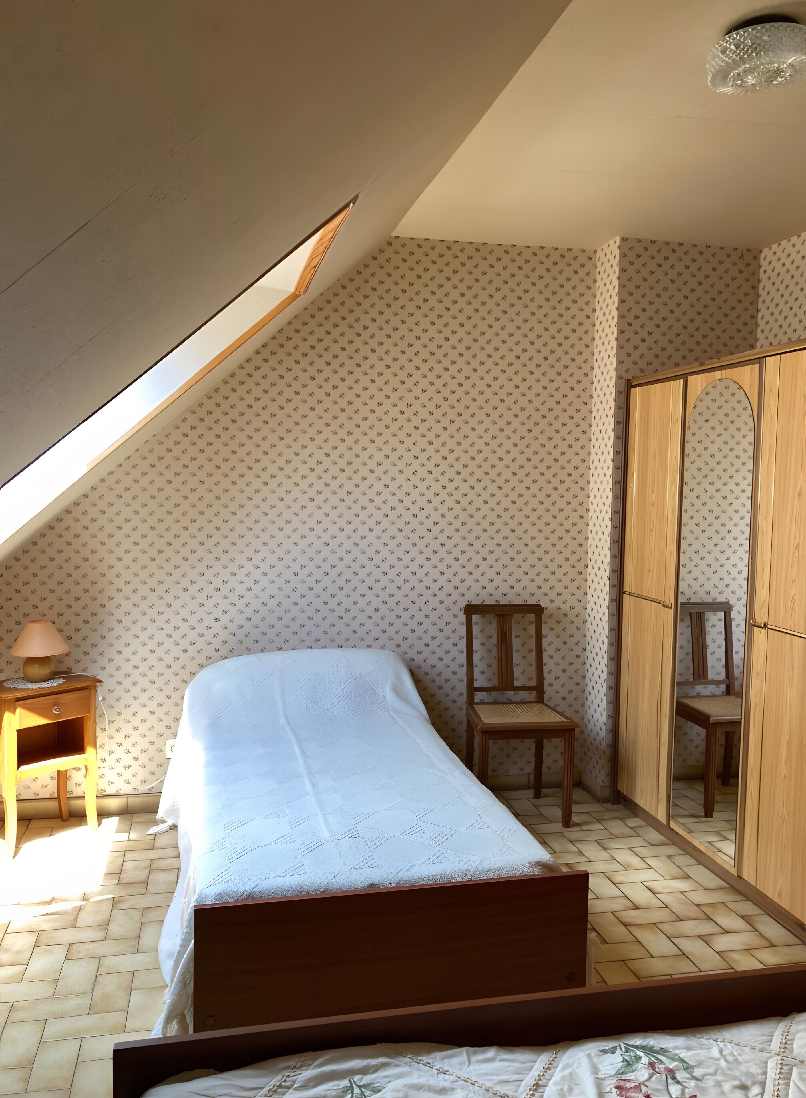 Un dormitorio con techo inclinado, una cama, una silla de madera, un armario y un papel tapiz estampado.