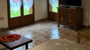 Huoneessa on televisio puisen kaapin päällä, koriste-esineillä koristeltu pöytä ja ikkuna, jossa on läpikuultavat verhot.