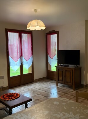 Une pièce avec un téléviseur posé sur un meuble en bois, une table ornée d’un objet décoratif et une fenêtre avec des rideaux légers.