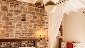 Un dormitorio con pared de piedra, una cama con cabecera, mesitas de noche y un rincón con cojines junto a la ventana.