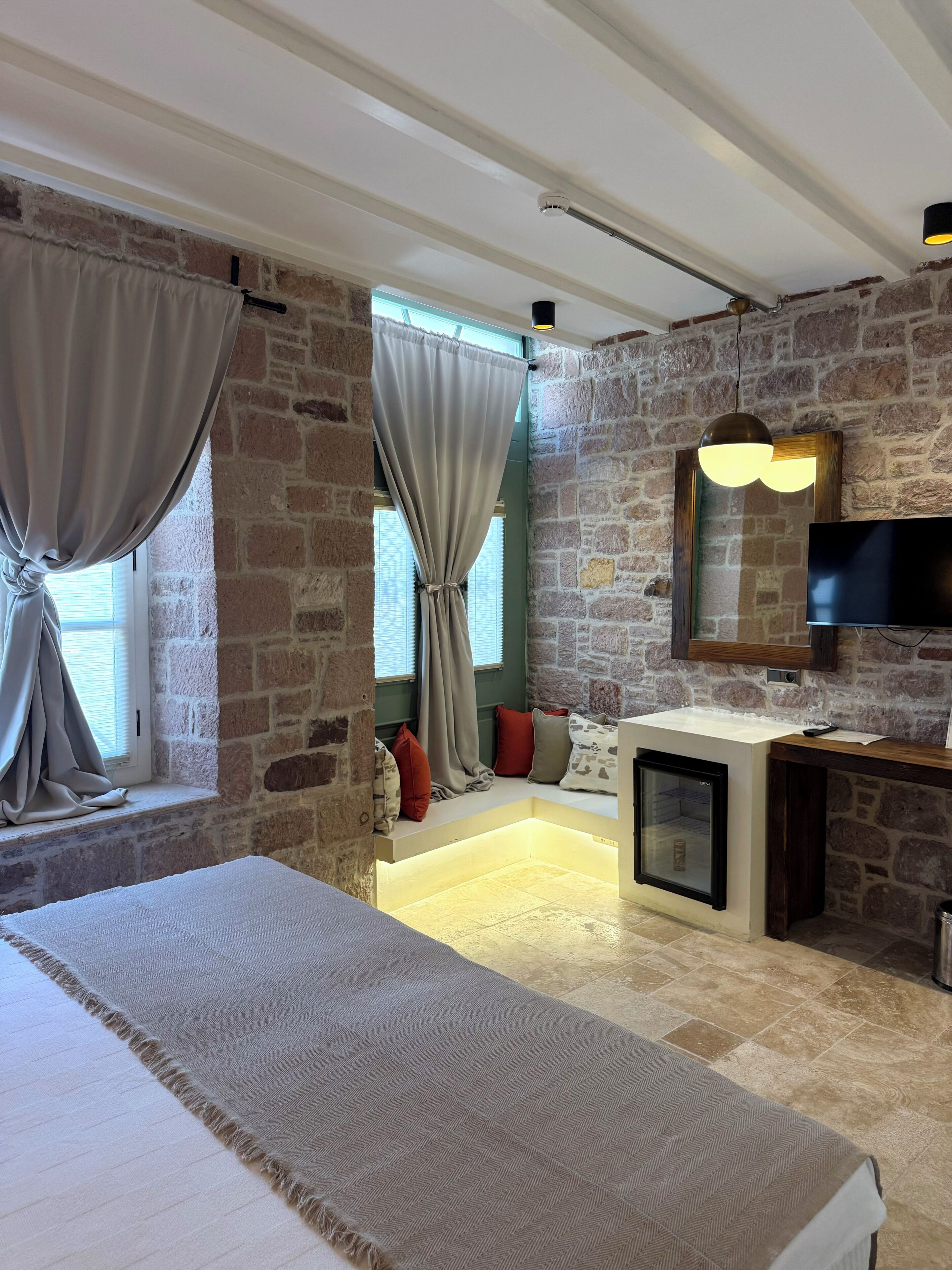 A bedroom with a stone wall, a bed, a TV, a small fridge, and a window with curtains.
