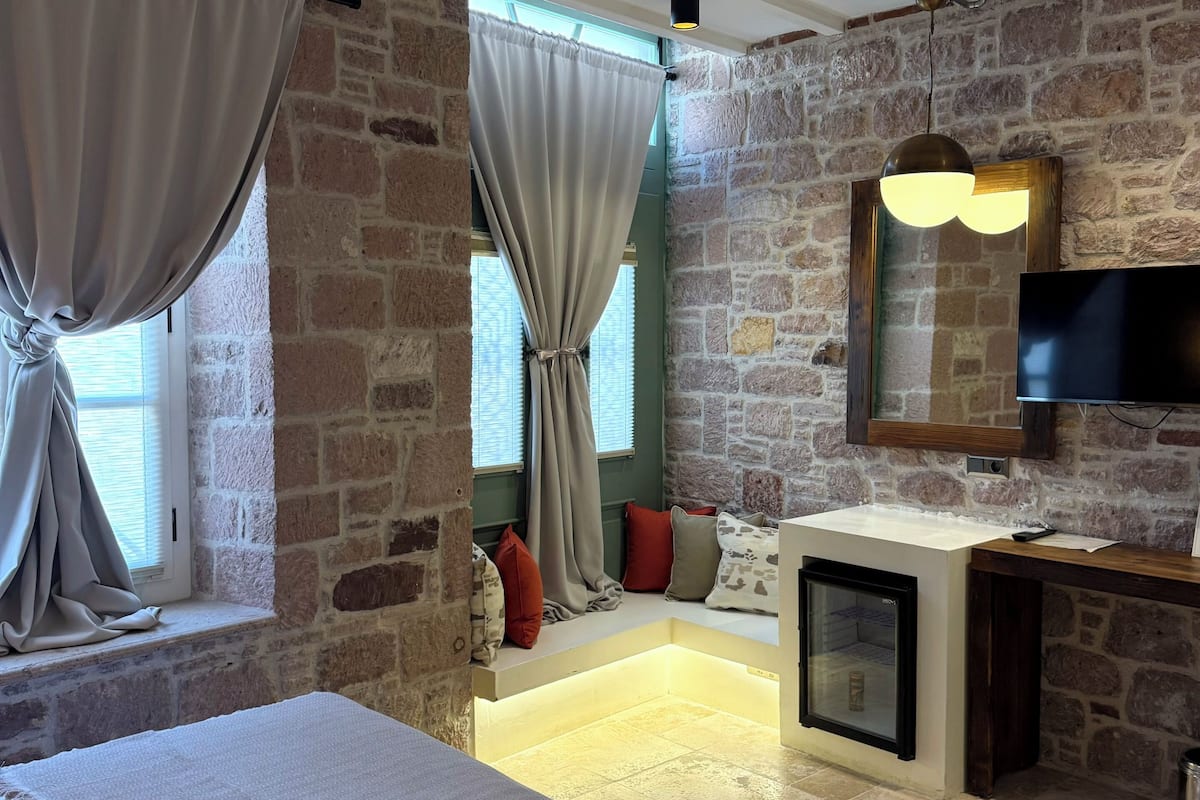 A bedroom with a stone wall, a bed, a TV, a small fridge, and a window with curtains.