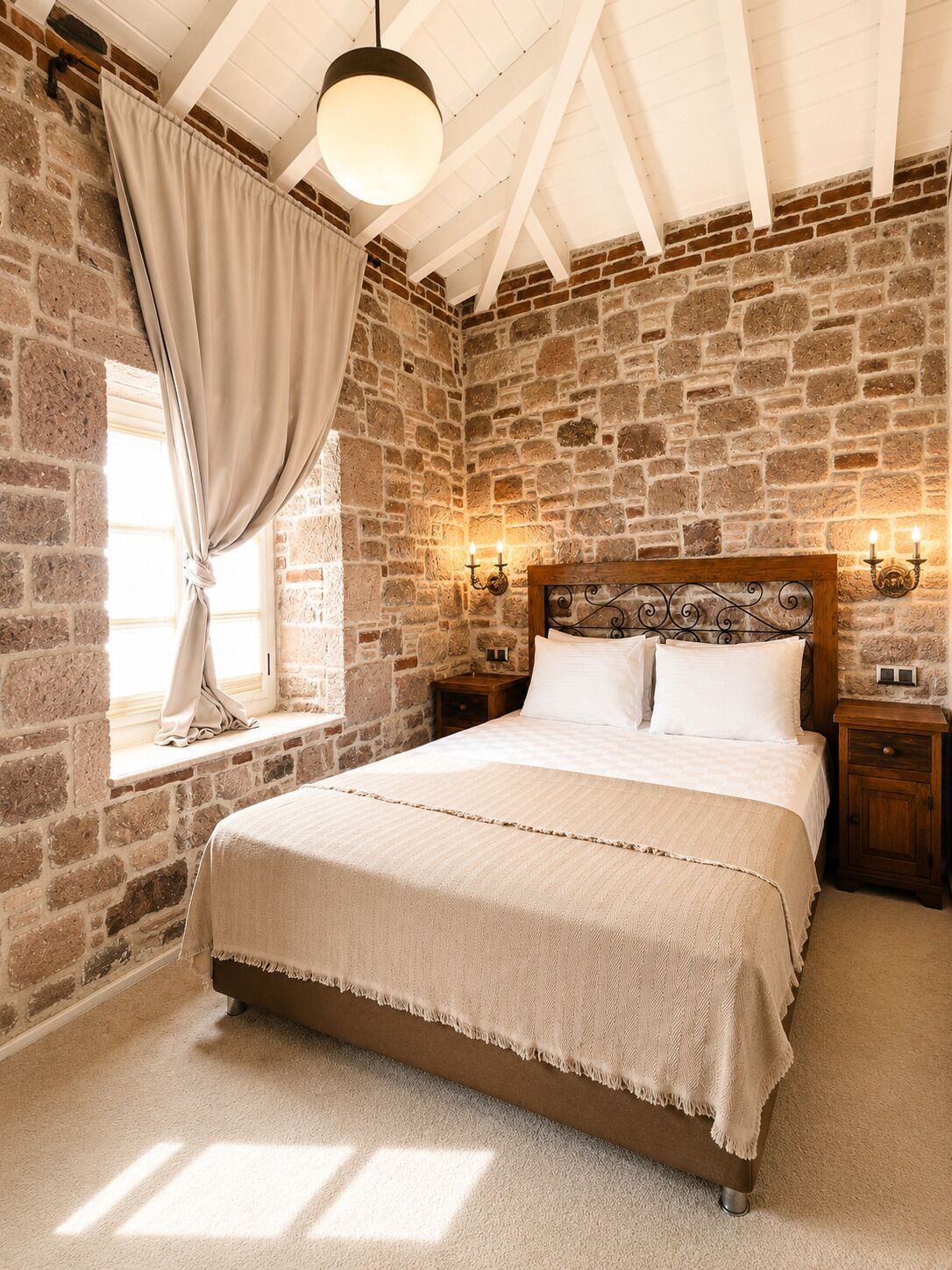 A bedroom with a stone wall, a large bed, a window with curtains, and wall-mounted lamps.