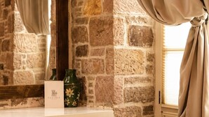 Una habitación con pared de piedra, un espejo, una ventana con cortinas, una estantería con adornos y un cuadro enmarcado.
