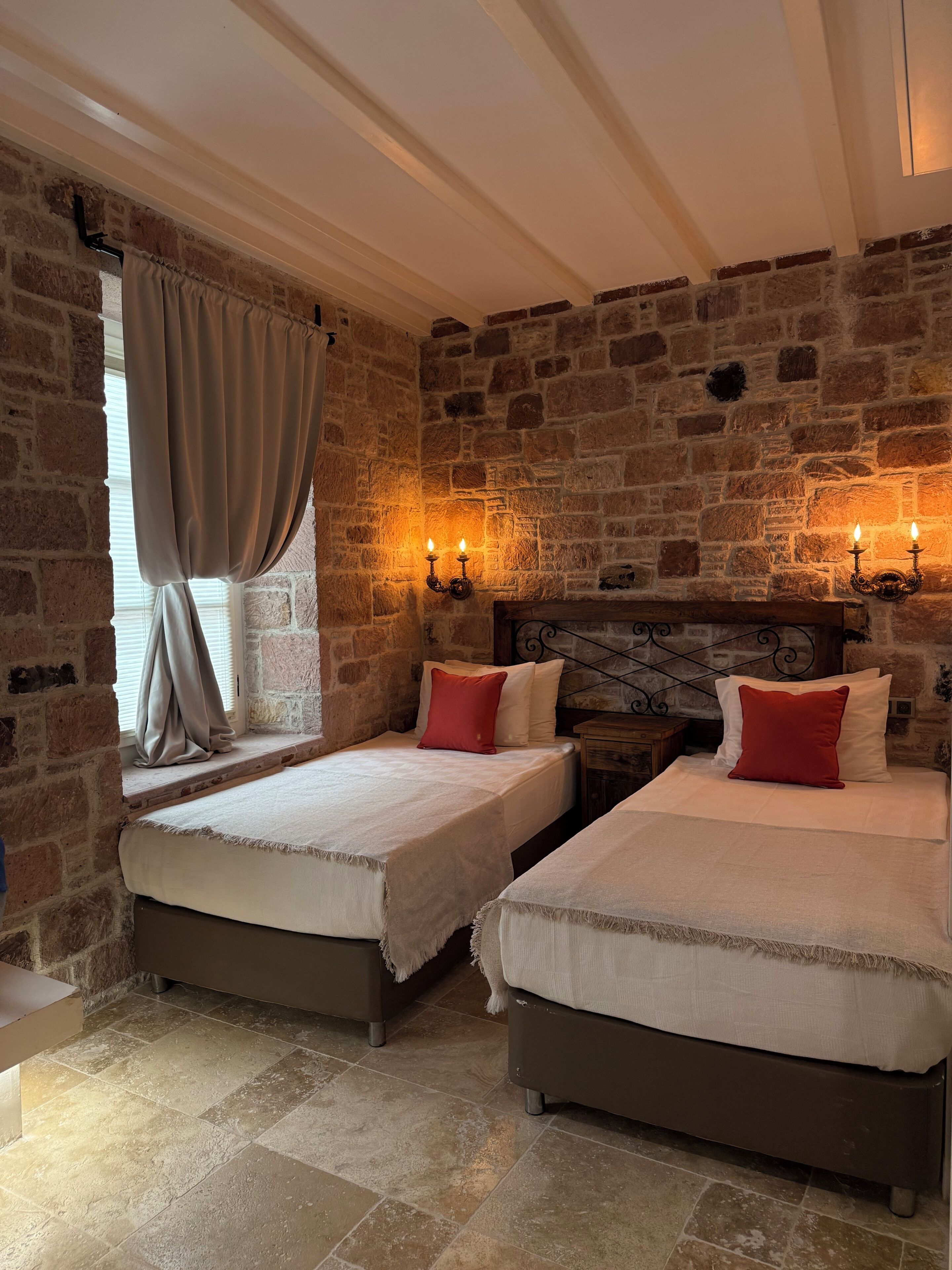 A room with two beds, stone walls, a window with curtains, and wall-mounted lights.