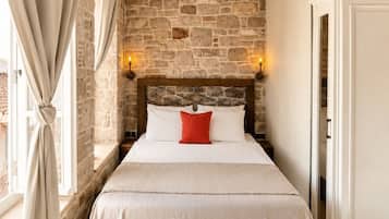 Une chambre avec un mur en pierre, un lit avec une literie blanche et un oreiller rouge, un luminaire suspendu et une porte sur la droite.