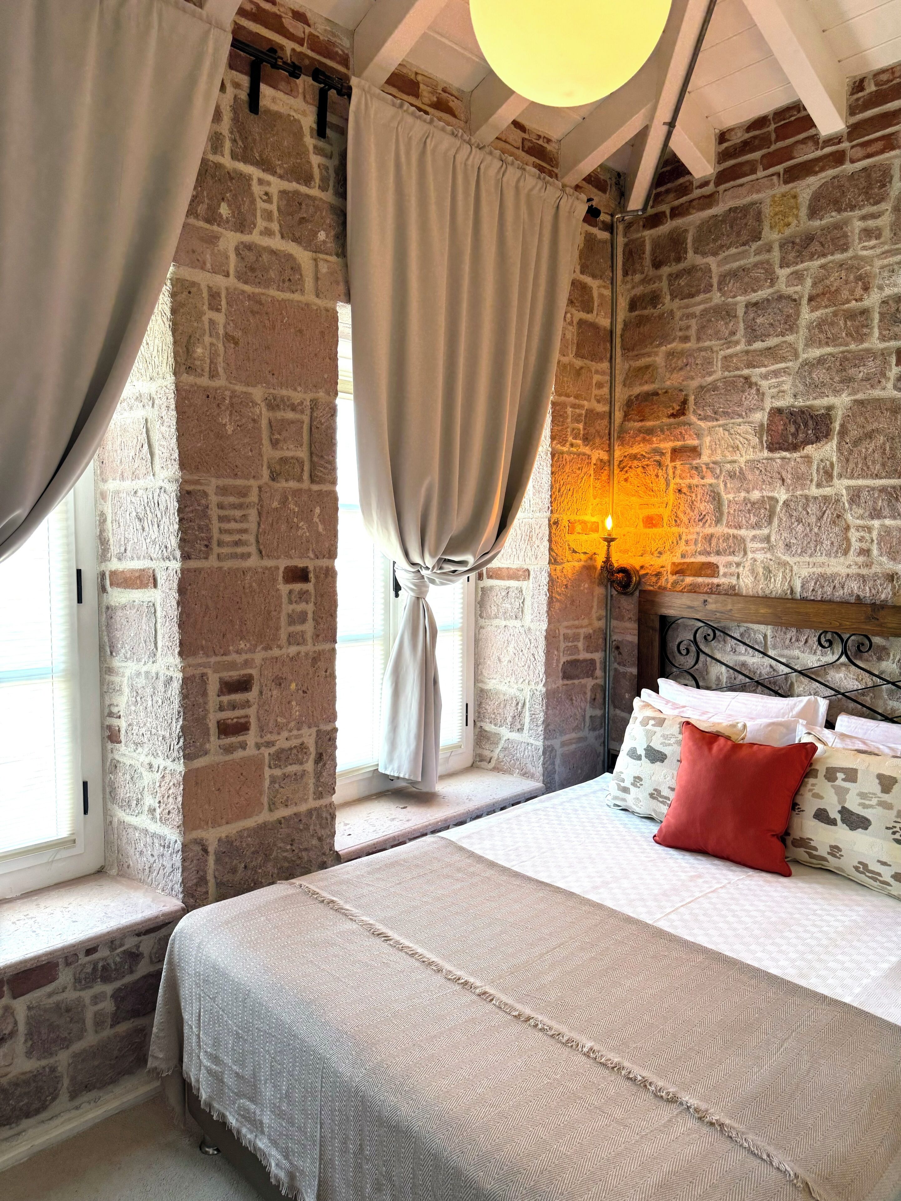 A bedroom with a stone wall, a bed with pillows, a window with curtains, and a hanging light fixture.