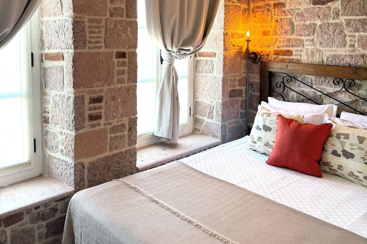 Ein Schlafzimmer mit Steinsandsteinwand, einem Bett mit Kissen, einem Fenster mit Vorhängen und einer Pendelleuchte.