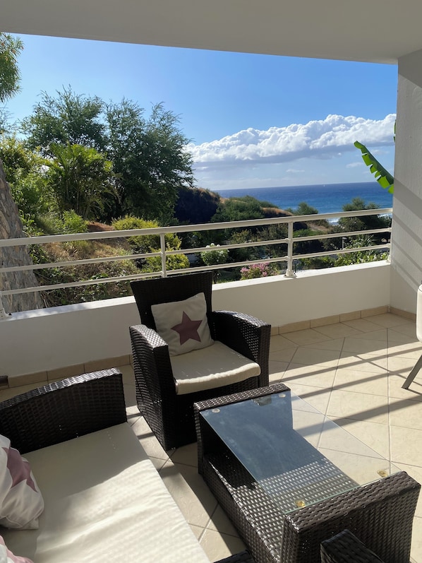 Ein Balkon mit Korbmöbeln, einem Glastisch und Blick aufs Meer.