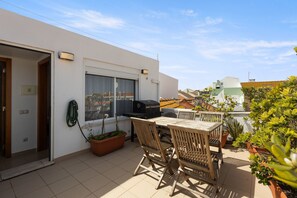 Una terraza en la azotea con una mesa, sillas, una parrilla y plantas en macetas.