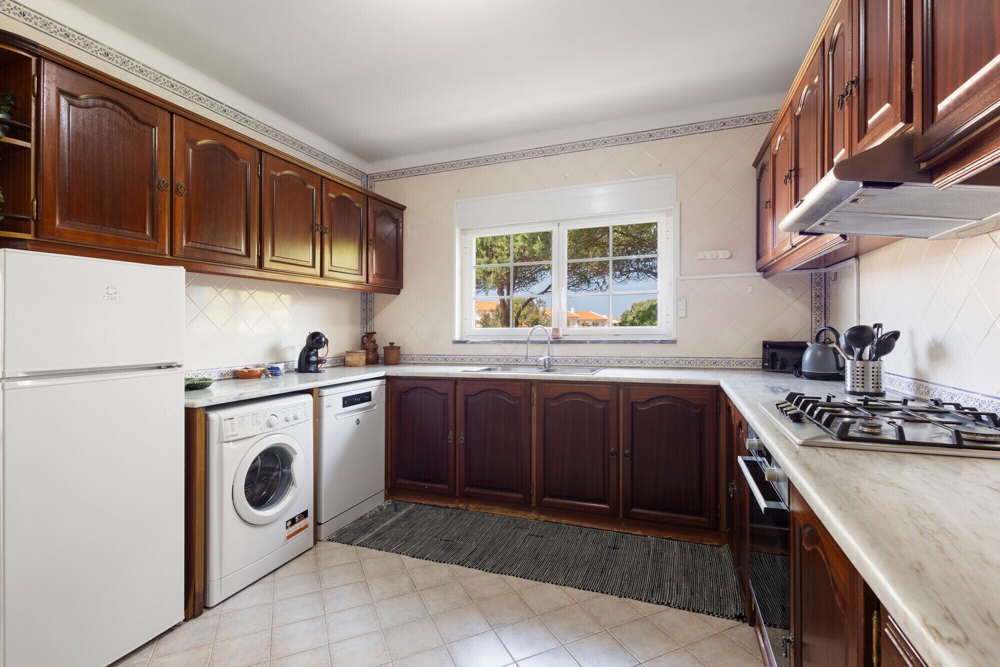 A kitchen with wooden cabinets, a built-in dishwasher, a stove, and a window with a view of trees.
