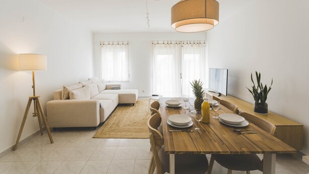 Ein modernes Wohnzimmer mit einem Holzesstisch, einem weißen Sofa und einem großen Fenster mit Vorhängen.