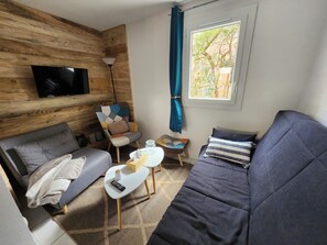 Ein modernes Wohnzimmer mit einem grauen Sofa, einem kleineren grauen Sessel, einem gemusterten Sessel, einem weißen Couchtisch mit einer Fernbedienung und einem an einer Holzwand befestigten Fernseher.