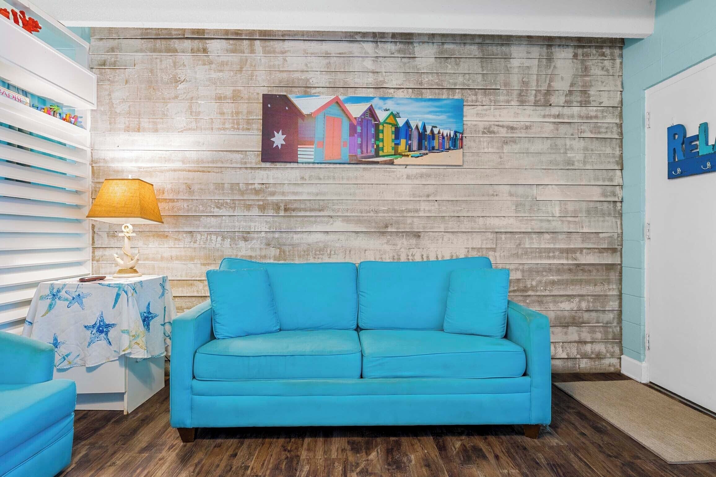 A room with a blue sofa, a wooden wall, a painting of colorful buildings, and a lamp.