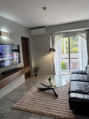 Ein modernes Wohnzimmer mit einem Flachbildfernseher, einem Glastisch, einer Ledersofa und Blick ins Freie durch ein großes Fenster.