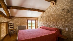A bedroom with a stone wall, wooden beams, a bed, a chair, and a window. - Holiday Home "La Cabane du Vigneron" with Shared Pool, Private Terrace & Air Conditioning (Donnazac)