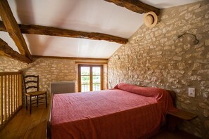 Ein Schlafzimmer mit Steinsandsteinwand, Holzbalken, einem Bett, einem Sessel und einem Fenster.