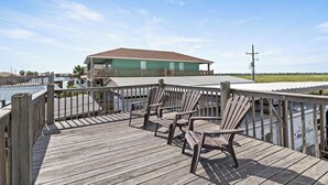 A wooden deck with Adirondack chairs, a building with a green roof, and a view of the water. - RV with AC on Bolivar Peninsula (Bolivar Peninsula)