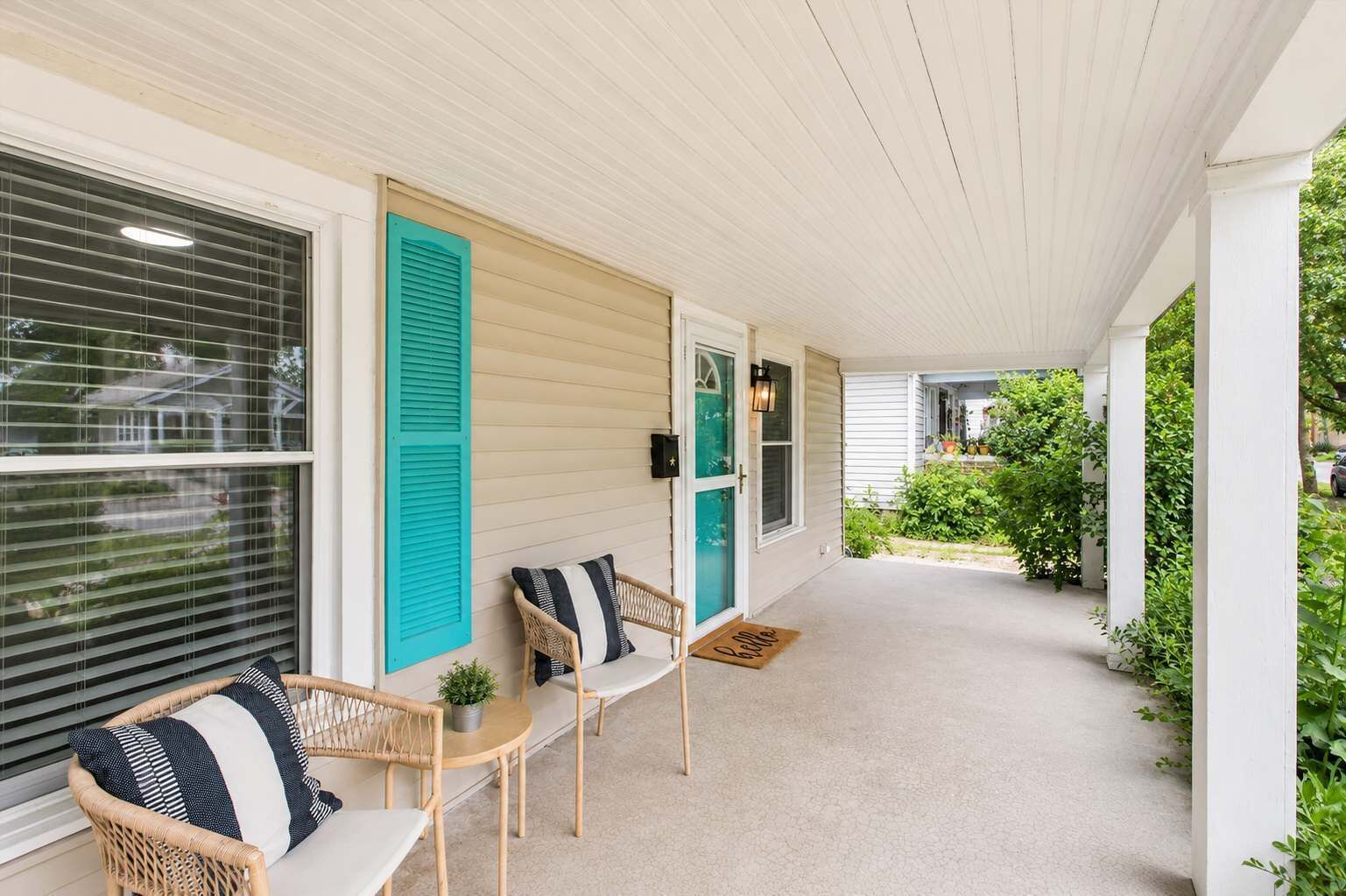 Une véranda couverte avec deux fauteuils en osier et une petite table, une porte avec un volet vert, et une vue sur un jardin.