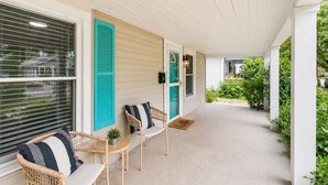 Eine überdachte Veranda mit zwei Korbstühlen und einem kleinen Tisch, eine Tür mit einem grünen Rollo und Blick auf einen Garten.