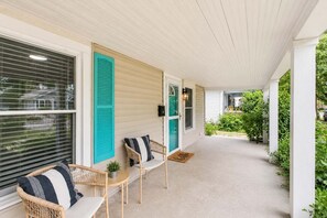 A covered porch with two wicker chairs and a small table, a door with a green shutter, and a view of a garden.