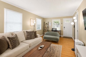 A living room with a sofa, armchair, coffee table, and a desk with a chair. There are two windows with blinds, a door, and a wall-mounted shelf with plants.