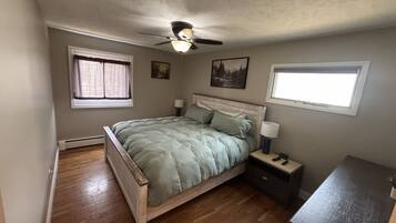 A bedroom with a bed, a nightstand, a desk, a ceiling fan, and two windows.