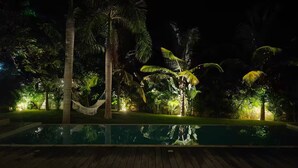 Ein Nachtblick auf einen Poolbereich mit Hängematte, Palmen und einer Holzterrasse.