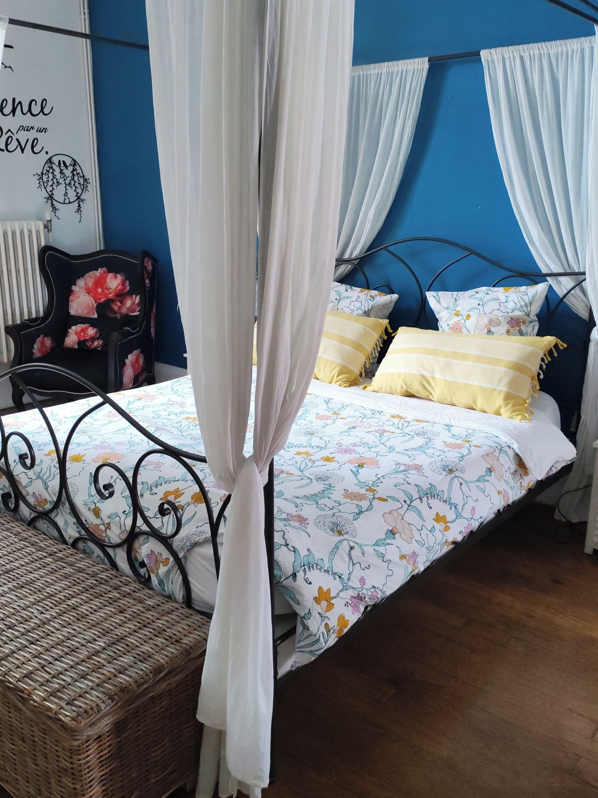 A bed with a floral bedspread, white curtains, and a wicker chair.