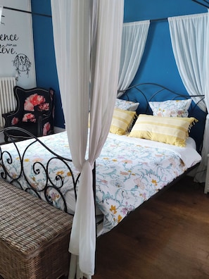Ein Bett mit floralem Überwurf, weißen Vorhängen und einem Korbstuhl.