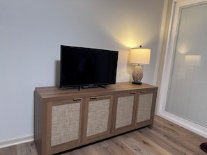 Un televisor sobre un mueble de madera con paneles trenzados, una lámpara y una ventana con persianas.