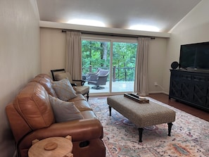 Una sala de estar con un sofá de cuero, un reposapiés estampado, un televisor sobre un mueble negro y vistas a un balcón con mobiliario exterior.