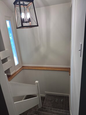 Una habitación de esquina con una silla blanca, un banco de madera, un ventanal pequeño y una lámpara colgante.