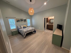 A bedroom with a bed, bedside tables, a hanging light fixture, and a view into the bathroom. - 3BR Beach Retreat – Short Walk to the Beach (Pine Knoll Shores)