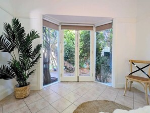 Ein Zimmer mit Blick zur Ecke, einer Zimmerpflanze, einem Sessel und einem Ausblick durch eine Glastür auf einen Außenbereich.