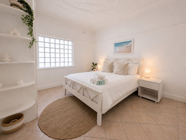 Une chambre à coucher blanche, avec un lit, des tables de chevet, une fenêtre et une étagère fixée au mur.