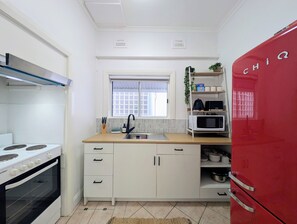 Une cuisine compacte avec des meubles blancs, une cuisinière, un évier, une fenêtre et un réfrigérateur rouge.