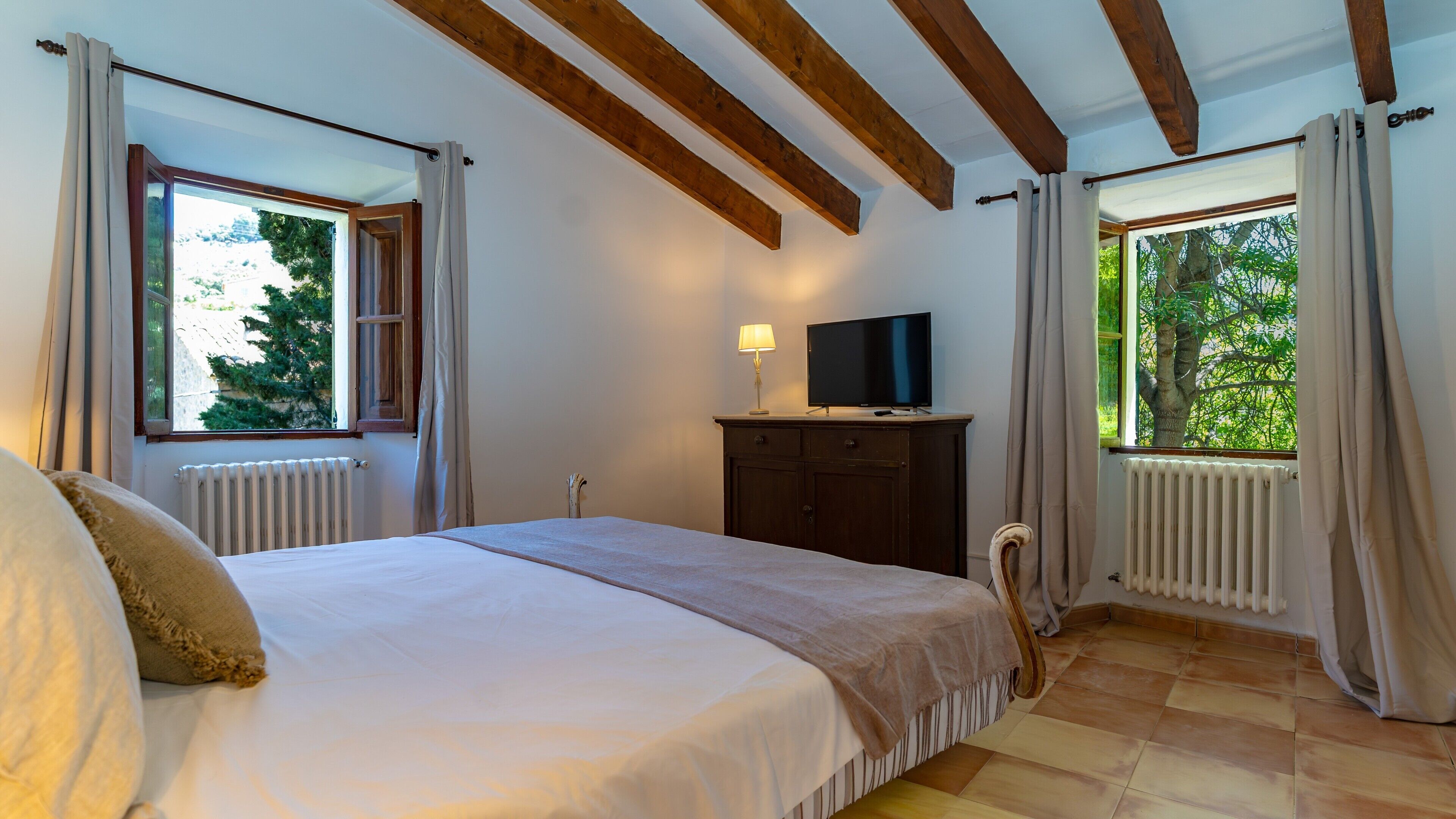 Un dormitorio con cama, televisor sobre un mueble y ventanas con cortinas.