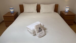 Ein ordentlich bezogenes Bett mit weißen Leinentüchern, zwei Kissen und ein gefaltetes Handtuch mit einem Logo auf einem Nachttisch aus Holz.