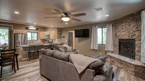 A spacious living room with a stone fireplace, a kitchen with wooden cabinets, and a ceiling fan. - Modern Cabin in Beautiful Pinetop (Pinetop-Lakeside)