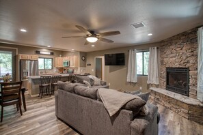 A spacious living room with a stone fireplace, a kitchen with wooden cabinets, and a ceiling fan. - Modern Cabin in Beautiful Pinetop (Pinetop-Lakeside)