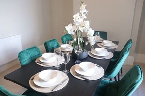 A dining table set with white plates, bowls, and cutlery, surrounded by teal chairs, with a vase of white flowers in the center.