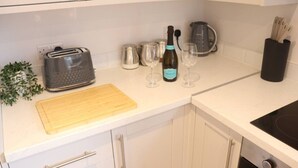 A kitchen with white cabinets, a toaster, a cutting board, a wine bottle, glasses, a kettle, and a black utensil holder. - Seaview Terrace - 5 Bedroom seaview home (Tyne and Wear)