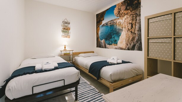 Ein Hotelzimmer mit zwei Betten, einem großen Gemälde einer Küstenlandschaft, einer Wandleuchte und einem Regal mit Aufbewahrungsboxen.