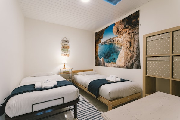 Ein Hotelzimmer mit zwei Betten, einem großen Gemälde einer Küstenlandschaft, einer Wandleuchte und einem Regal mit Aufbewahrungsboxen.