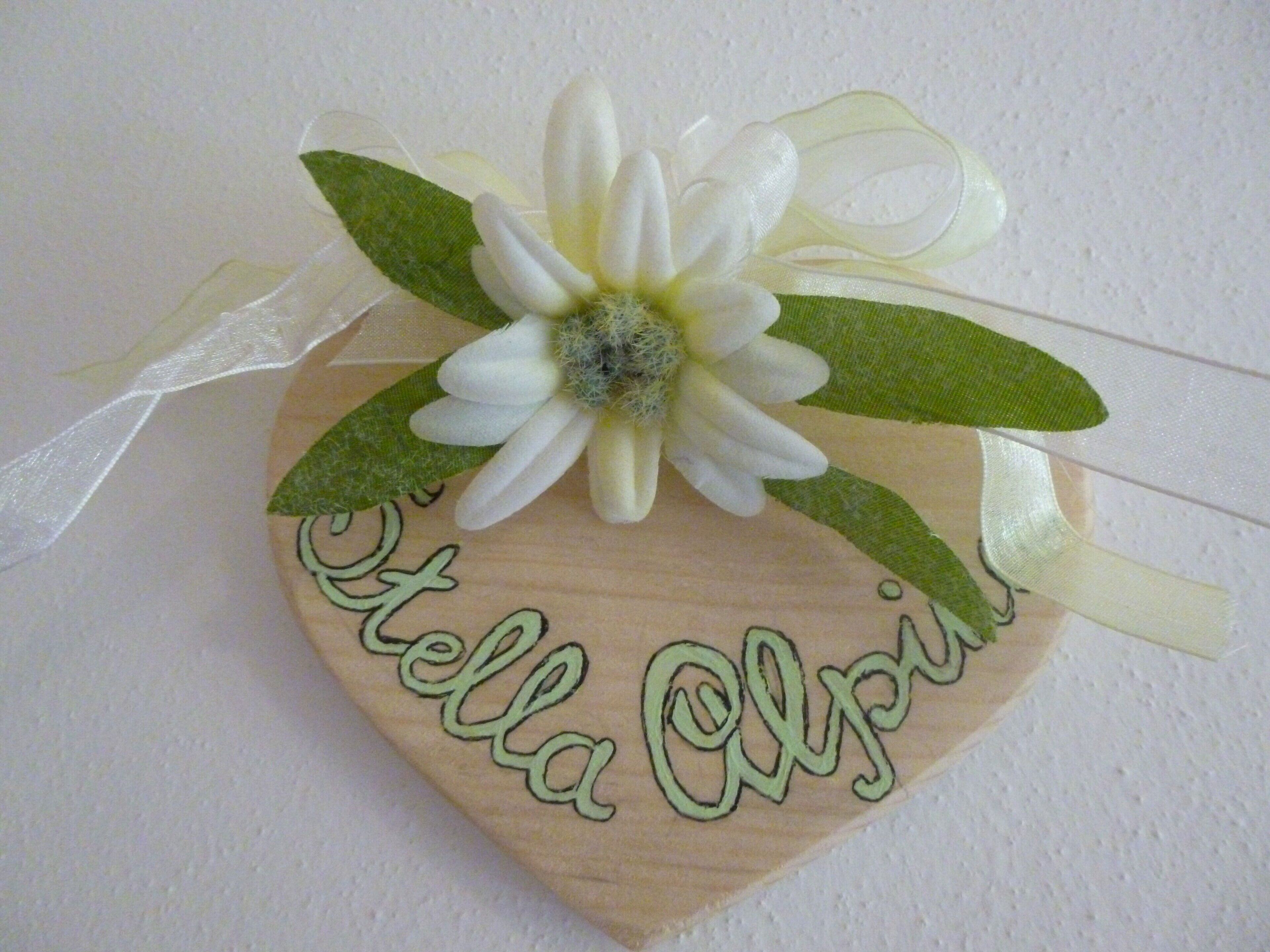 A wooden heart-shaped decoration with a white flower and green leaves, featuring the name 'Stella Olivia' embroidered in green.