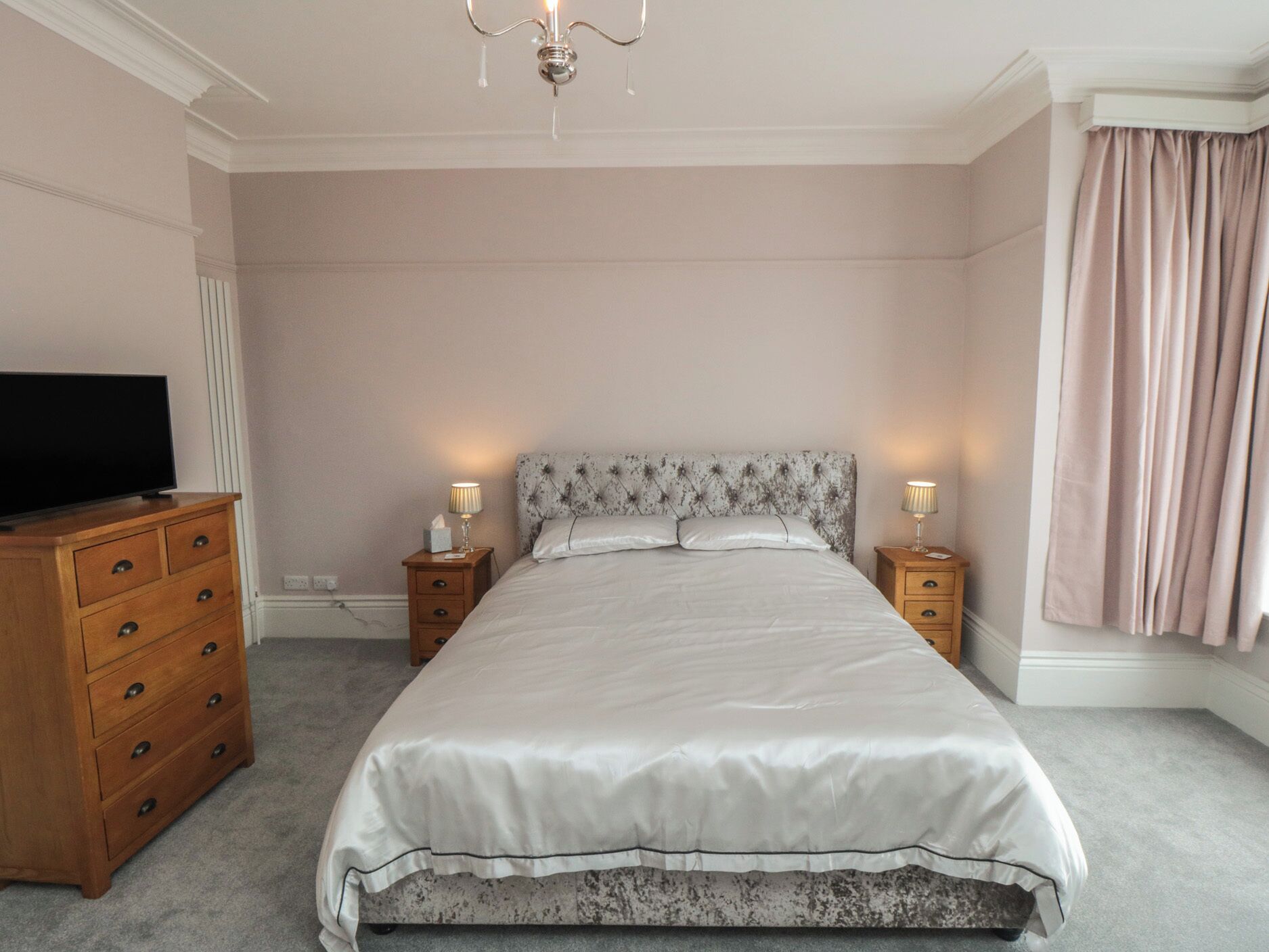 Een slaapkamer met een bed, een televisie op een dressoir, twee bedlampen en een kroonluchter.