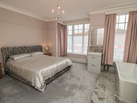 Un dormitorio con cama, mesita de noche, candelabro, ventana con cortinas, baño con lavamanos y espejo, y bañera.