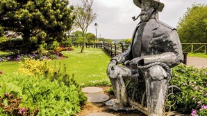 A statue of a seated figure with a pipe, surrounded by a garden with various plants and a fence in the background. - Ruby's Retreat (Filey)