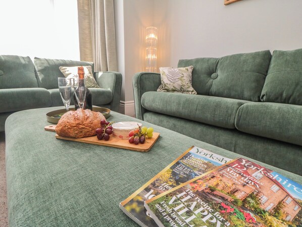 Un salon avec un canapé vert, une table en bois sur laquelle se trouvent une miche de pain, des raisins et une bouteille de vin, ainsi qu’un magazine posé sur la table.